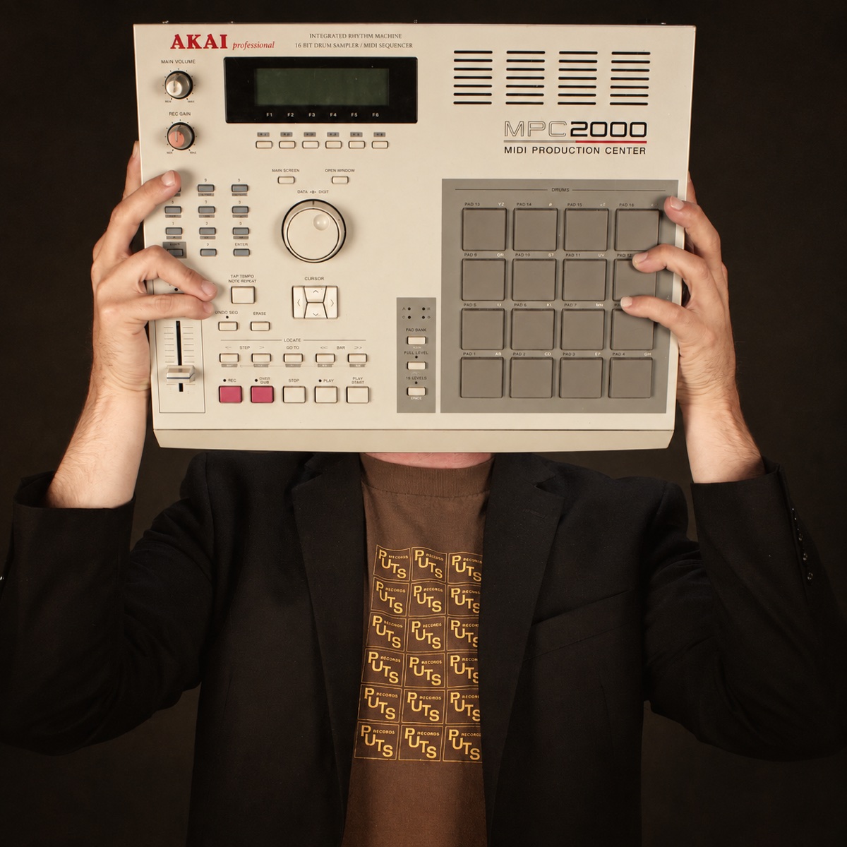 Tizz Tarkington holding an Akai MPC2000 sampler in front of his face, wearing a black blazer.